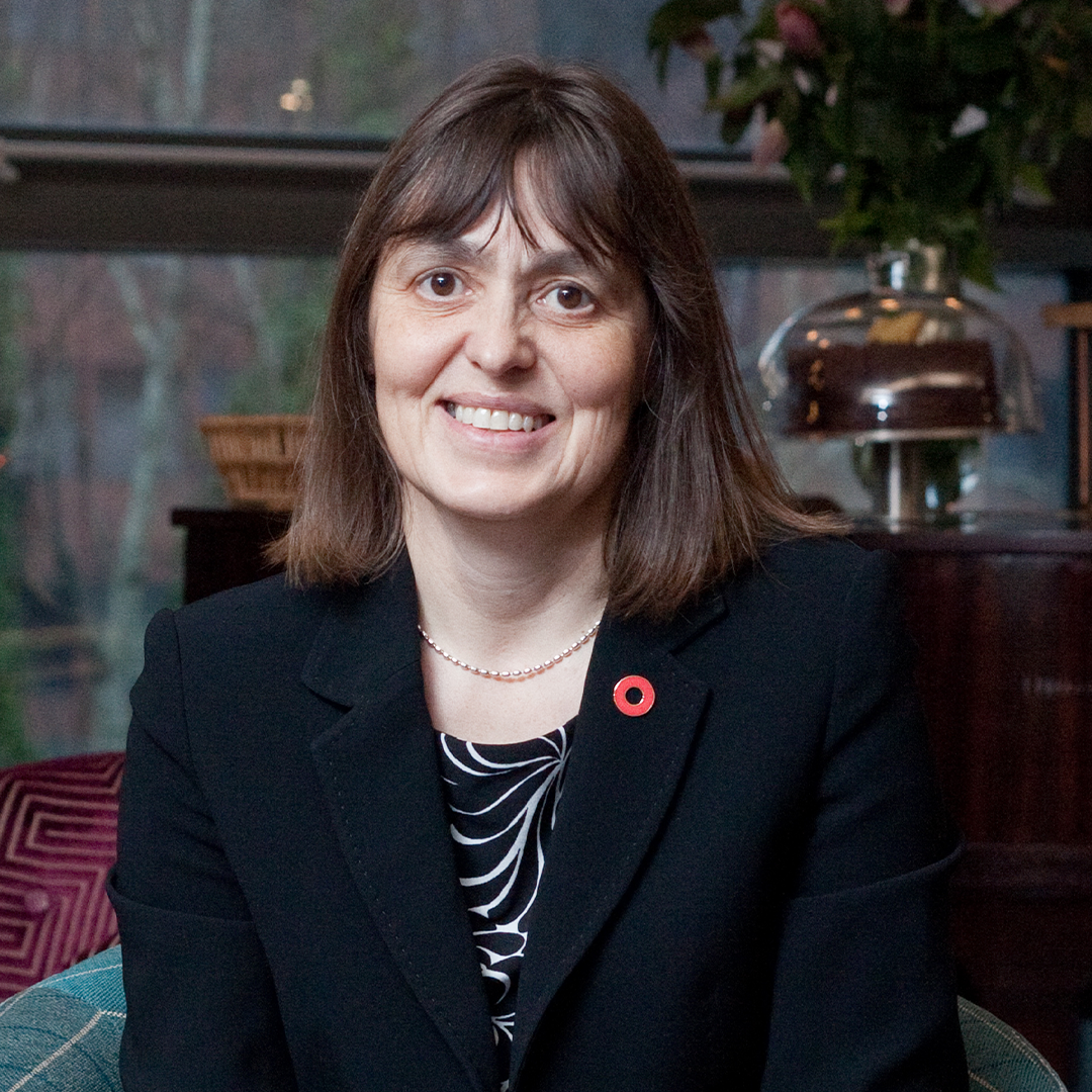 A person dressed in a black blazer with a patterned top underneath, seated indoors. The blazer has a red poppy pin on the lapel, and the individual is wearing a pearl necklace. The background features a glass window, a plant arrangement, and decorative items on a wooden surface.