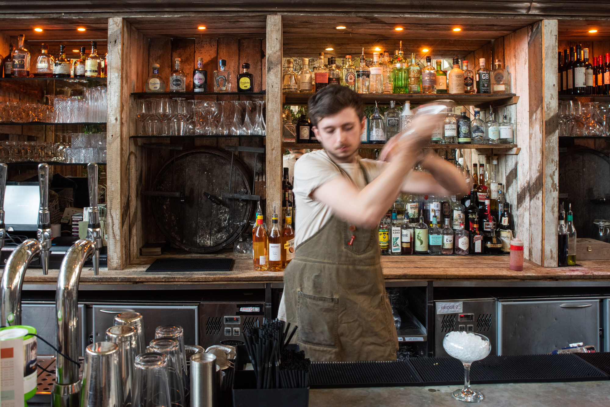 Bartender behind a rustic wooden bar shaking a cocktail shaker. The bar is stocked with various bottles of spirits, wine, and mixers arranged on shelves, along with glassware and a large wooden barrel in the center. The countertop has cocktail-making tools, straws, and an empty glass with ice ready for a drink. Warm lighting creates a cozy atmosphere.