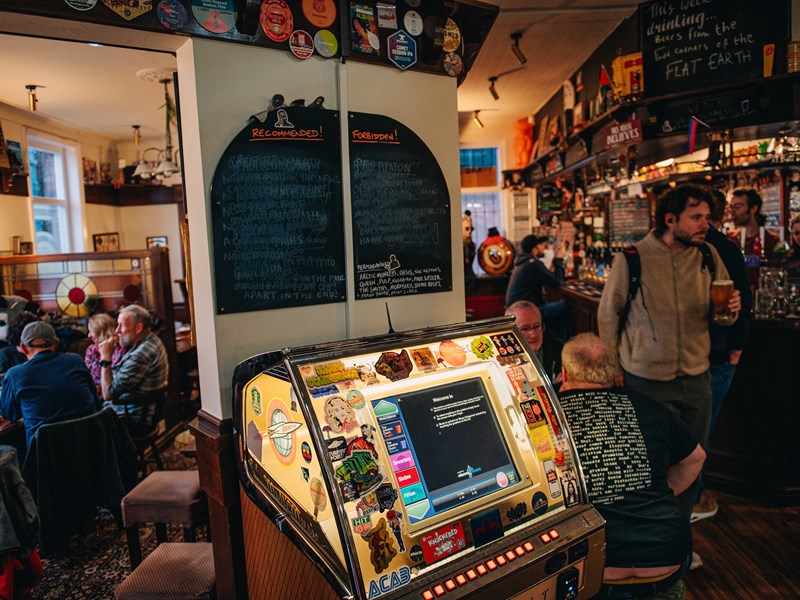 Rutland Arms jukebox lit up, with a chalk board above that's separated into forbidden and recommended. Groups of people are sat to the left of the jukebox, with the bar to the right.