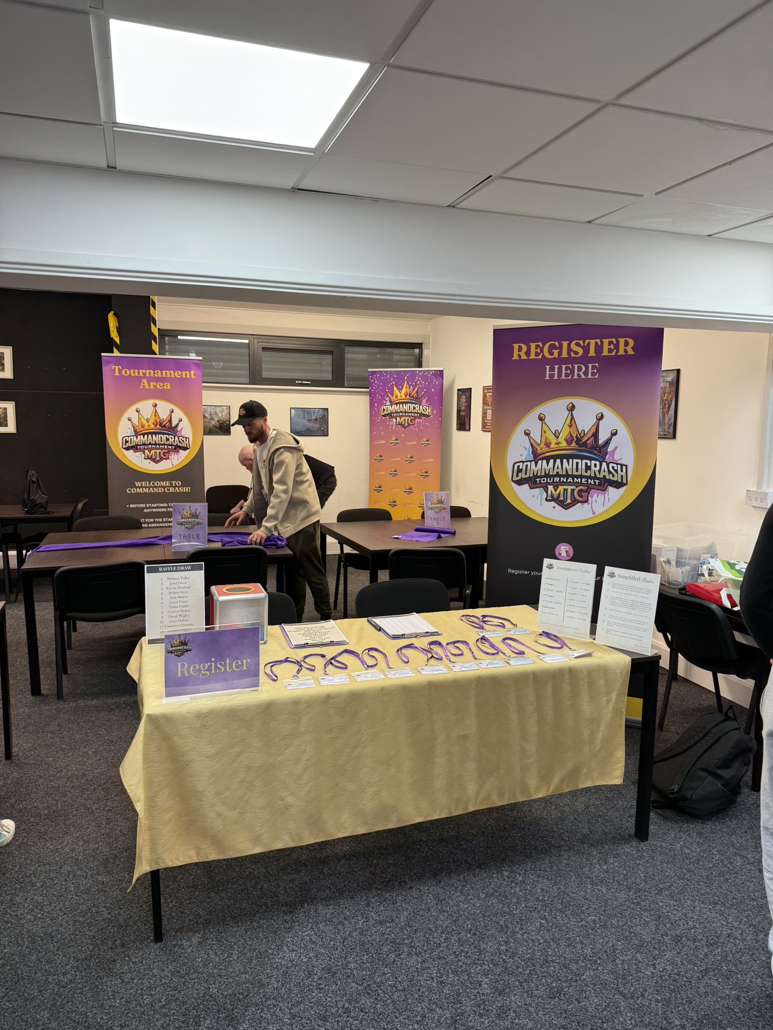 A registration area for the “Command Crash MTG Tournament” featuring tables covered with gold and purple cloths. The front table displays registration forms, lanyards, and signs with event details. Behind it are two large banners: one reads “Register Here” with the tournament logo, and another marks the “Tournament Area.” A person is arranging items on a back table. The room has framed artwork on the walls and chairs around the tables.