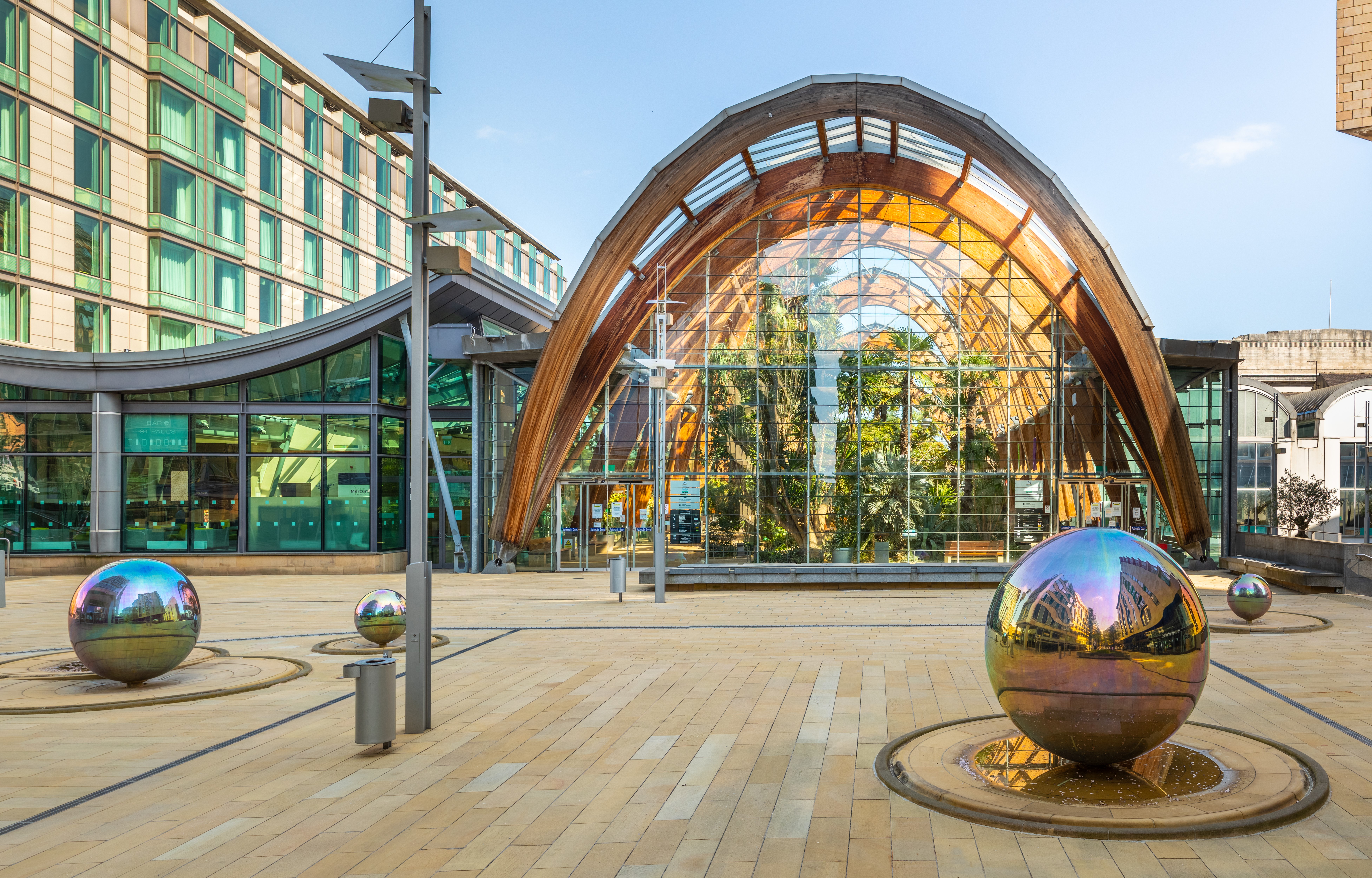 Winter Garden - Exterior view of the Winter Garden, featuring a large arched glass and timber structure with lush greenery visible inside. In the foreground, a paved courtyard displays several reflective stainless steel spheres set in shallow water features.