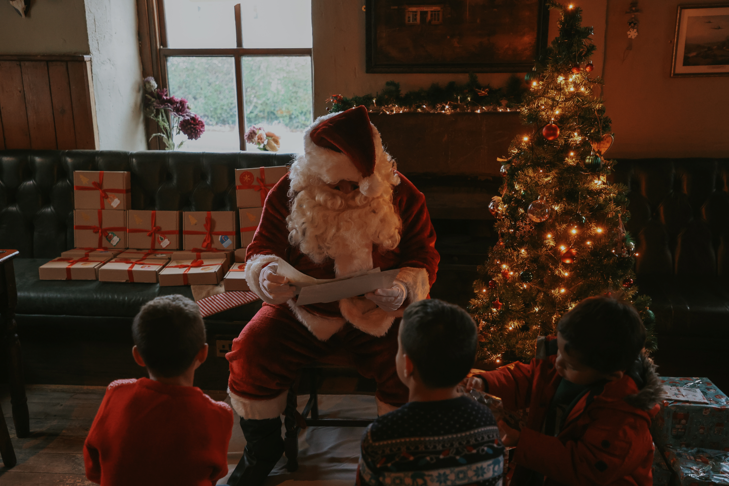 Santa Claus sits on a chair in a cozy, dimly lit room decorated for Christmas, reading a letter to three children seated on the floor in front of him. A decorated Christmas tree with glowing lights stands to the right, and wrapped presents are stacked on a bench to the left. A window and festive garlands add to the warm holiday atmosphere.