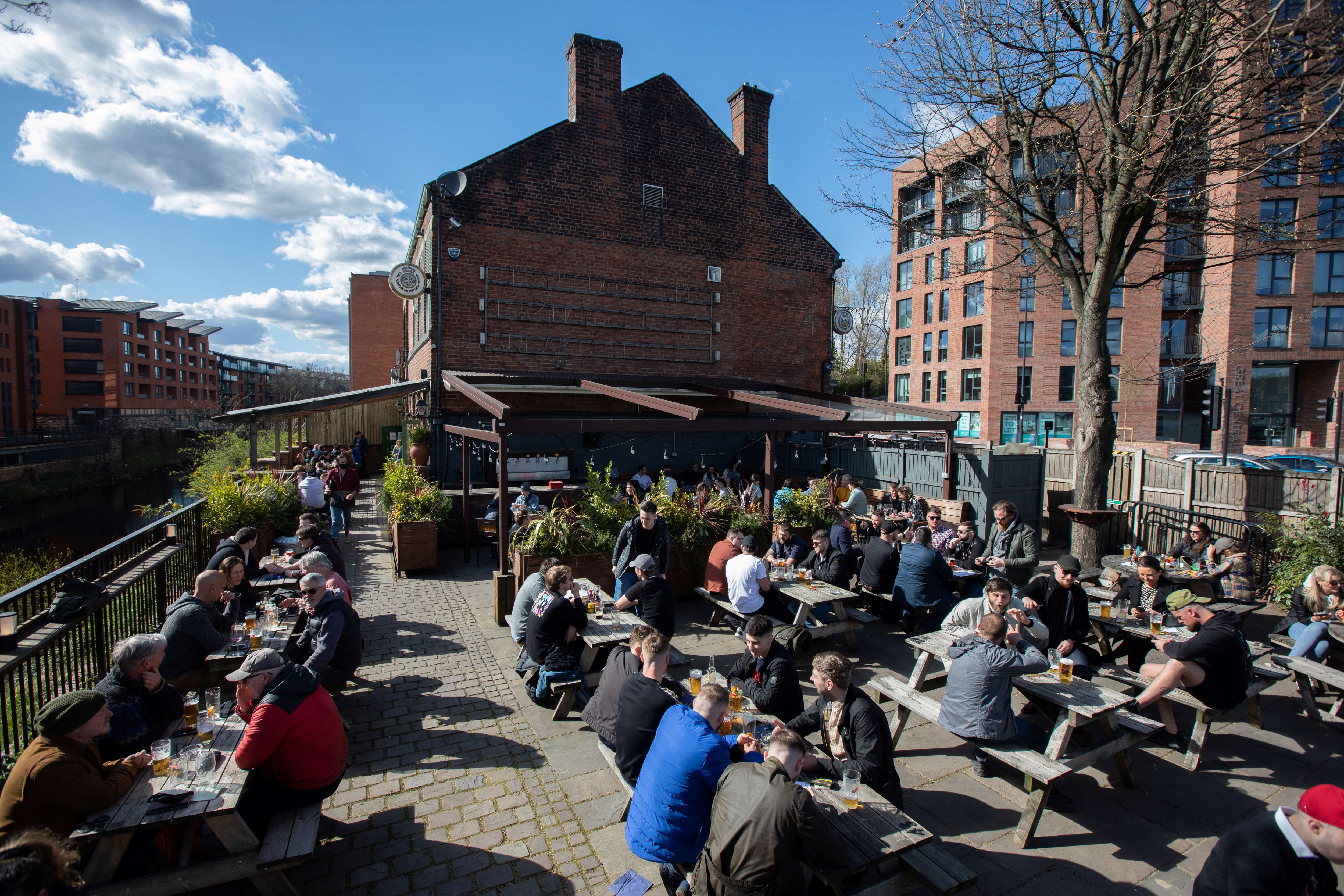 Outdoor seating area of a riverside pub filled with people at wooden picnic tables enjoying drinks in sunny weather. The space is paved with stone and lined with planters, next to a canal with railings. Behind the seating area is a large brick building with a pitched roof, and modern apartment blocks rise in the background under a bright blue sky with scattered clouds.