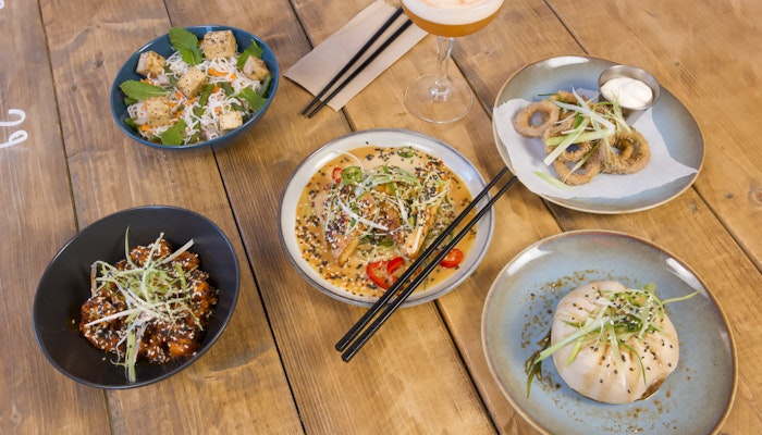 Overhead view of an assortment of Asian-inspired dishes arranged on a wooden table. The spread includes a steamed bun topped with sliced green onions, a plate of crispy fried rings with dipping sauce, a bowl of glazed meat garnished with sesame seeds, a dish of tofu cubes with herbs, and a plate of dumplings in a creamy sauce sprinkled with sesame seeds and sliced chili. Black chopsticks rest on two plates, and a cocktail in a coupe glass is placed near the center.