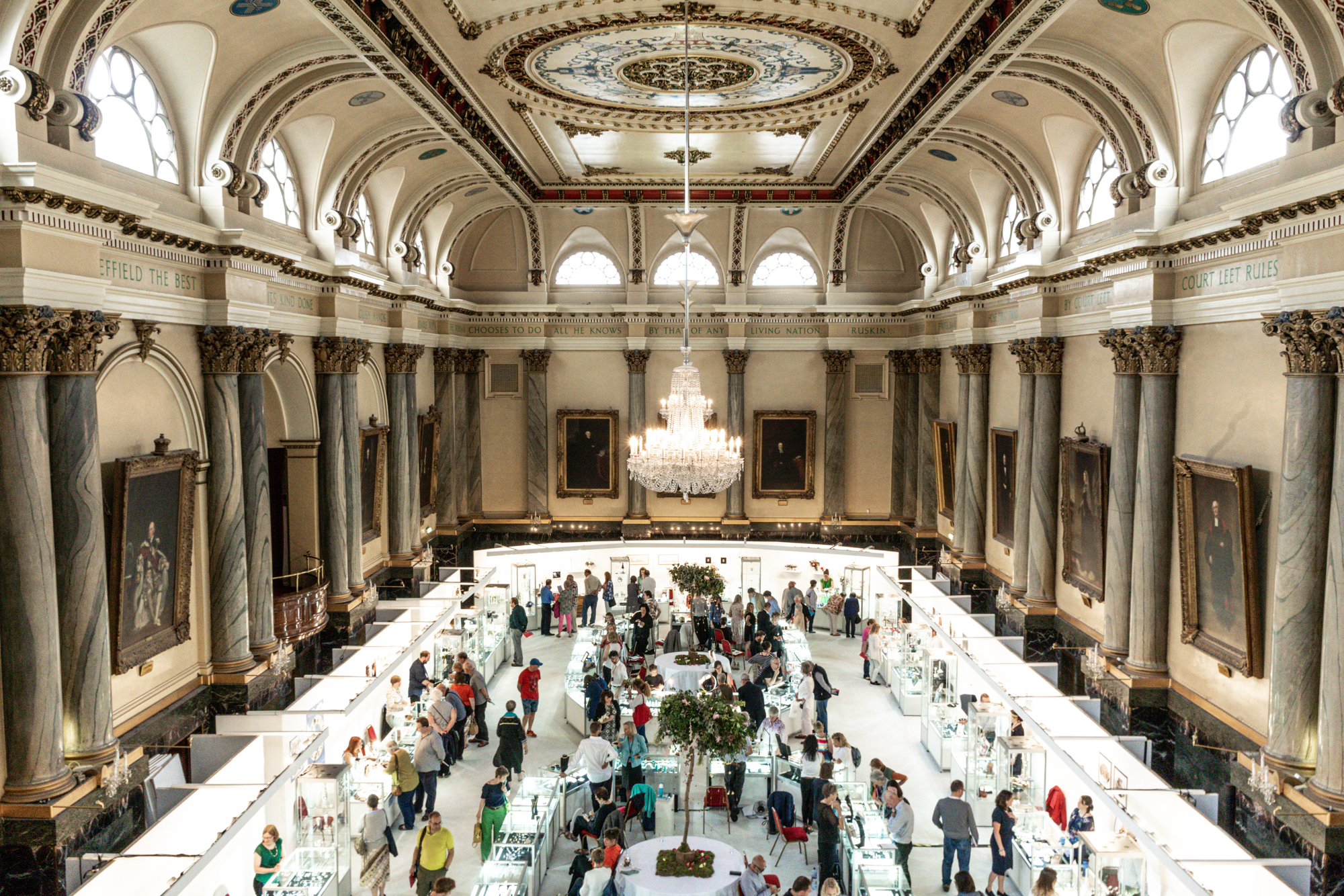 Elegant exhibition inside a grand hall with ornate columns, arched windows, and a decorative ceiling featuring a large chandelier. Visitors browse display booths arranged in a square layout around a central tree.