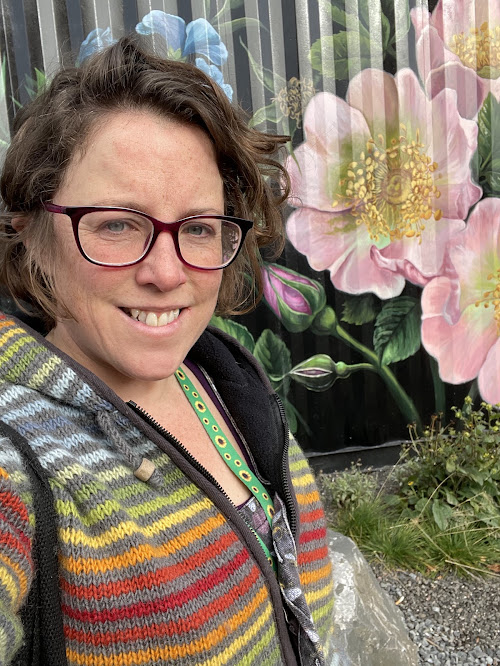 A person wearing a colorful striped wool cardigan with shades of red, yellow, green, and gray, standing in front of a mural featuring large, detailed pink and blue flowers painted on a dark background. The scene includes gravel and greenery at the bottom of the mural.