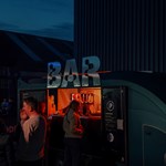 Outside 'bar' truck at Peddler Market. A woman is waiting at the truck and there are groups of people socialising nearby.