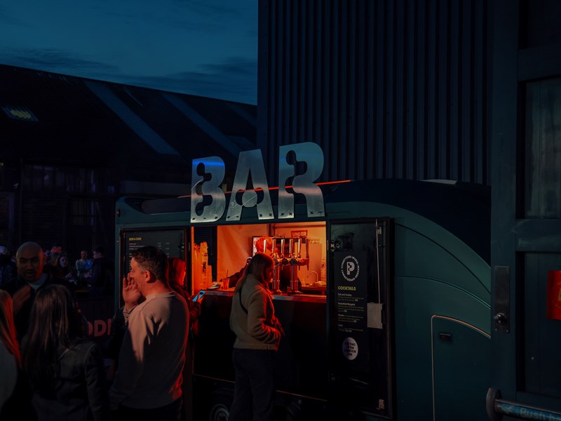 Outside 'bar' truck at Peddler Market. A woman is waiting at the truck and there are groups of people socialising nearby.